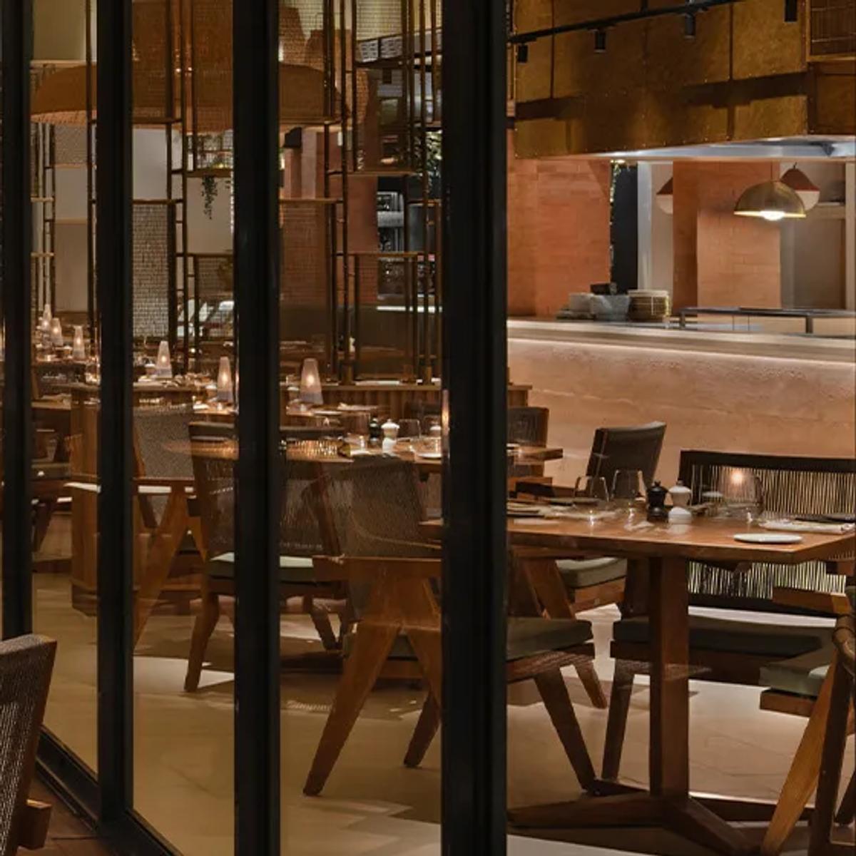The Citronelle Club's dining room at SO/ Maldives at night, candlelit tables lined along a glass wall beside the open exhibition kitchen