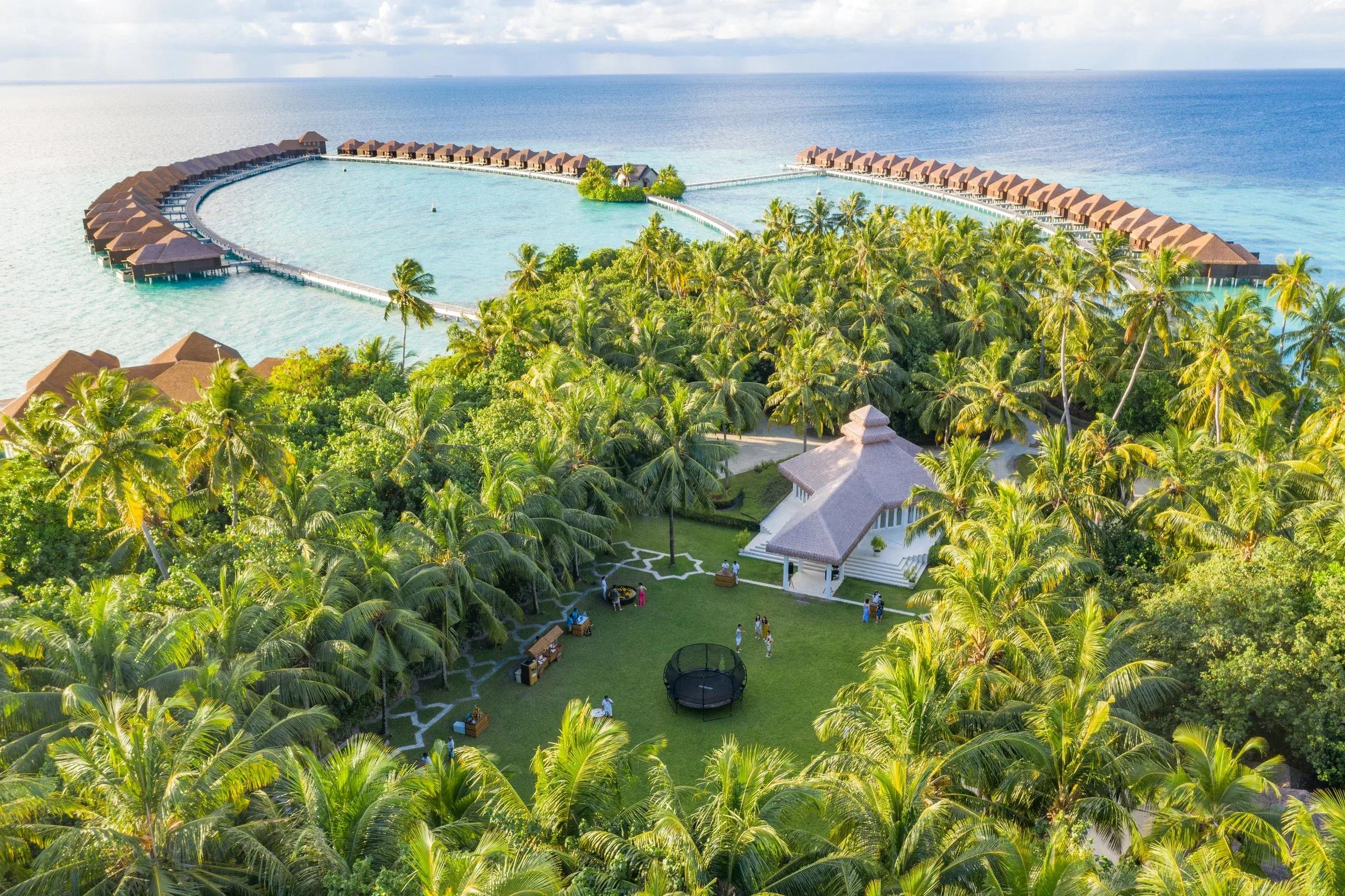 Aerial view of Ayada Maldives — the horseshoe of overwater villas enclosing a shallow lagoon, with the garden and lawn pavilion in the foreground