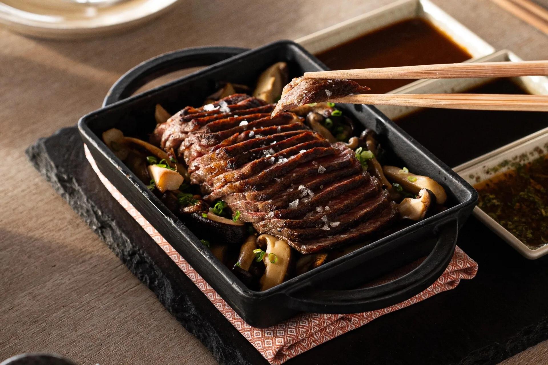 A sliced, seared dish lifted with chopsticks from a hot cast-iron pan at Kayto, dipping sauces on the side