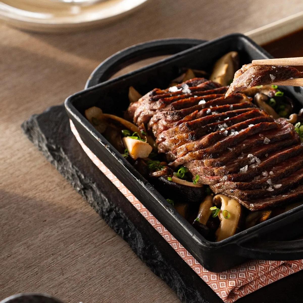 A sliced, seared dish lifted with chopsticks from a hot cast-iron pan at Kayto, dipping sauces on the side