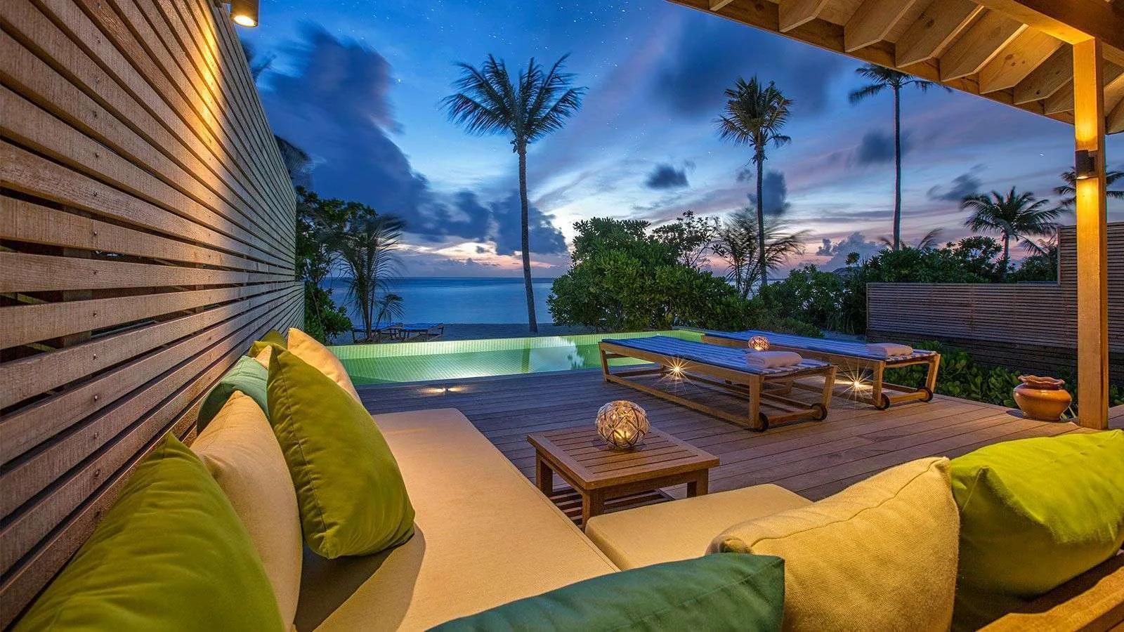A Beach Sunset Pool Villa's infinity pool and deck lounge at dusk, palm trees silhouetted against the fading sky