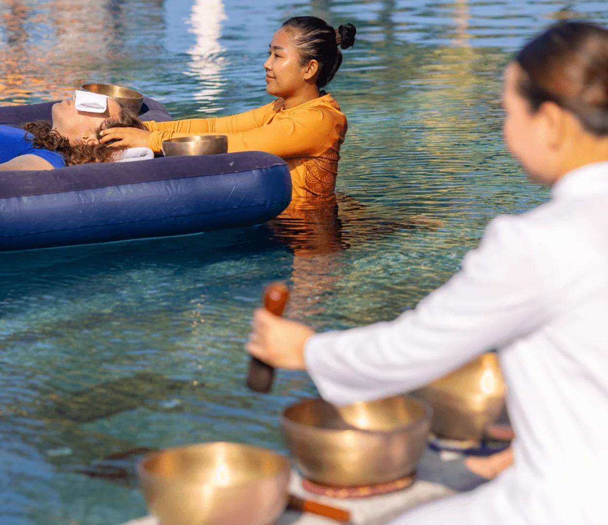 A guest receiving Floating Sound Healing at OBLU SELECT Lobigili, resting on an inflatable float while a therapist plays singing bowls in the water