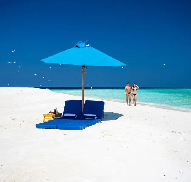 A blue sun umbrella and two loungers set up alone on a bare white sandbank, a couple walking the shoreline in the distance