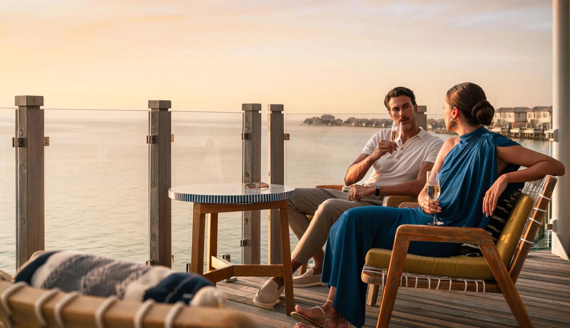 A couple sharing champagne on an overwater deck at sunset, with villas lined up behind them