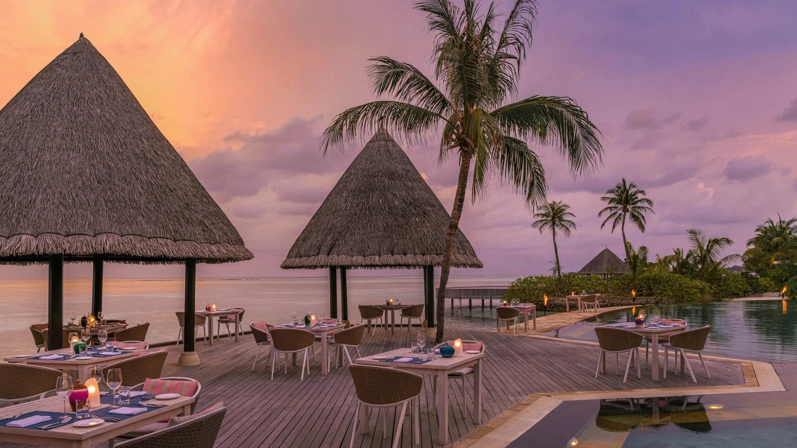 Kandu Grill's thatched pavilions lit at dusk, tables set beside the pool and lagoon