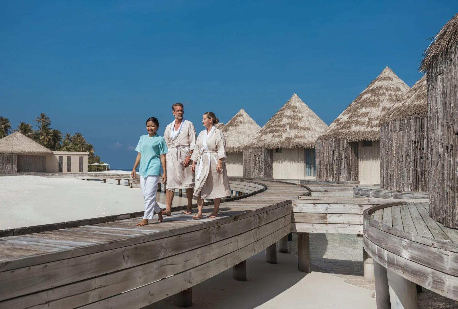 A couple in robes led by a therapist along the boardwalk to AVI Spa's overwater treatment villas