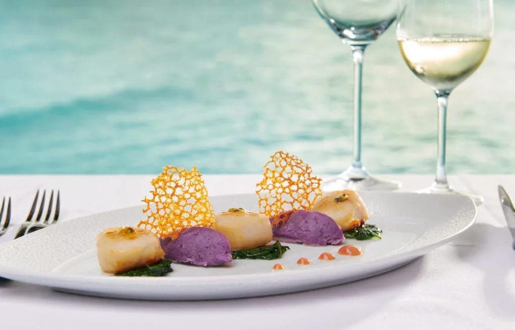 A plated seafood dish with scallops and purple potato purée, two glasses of white wine and the ocean behind