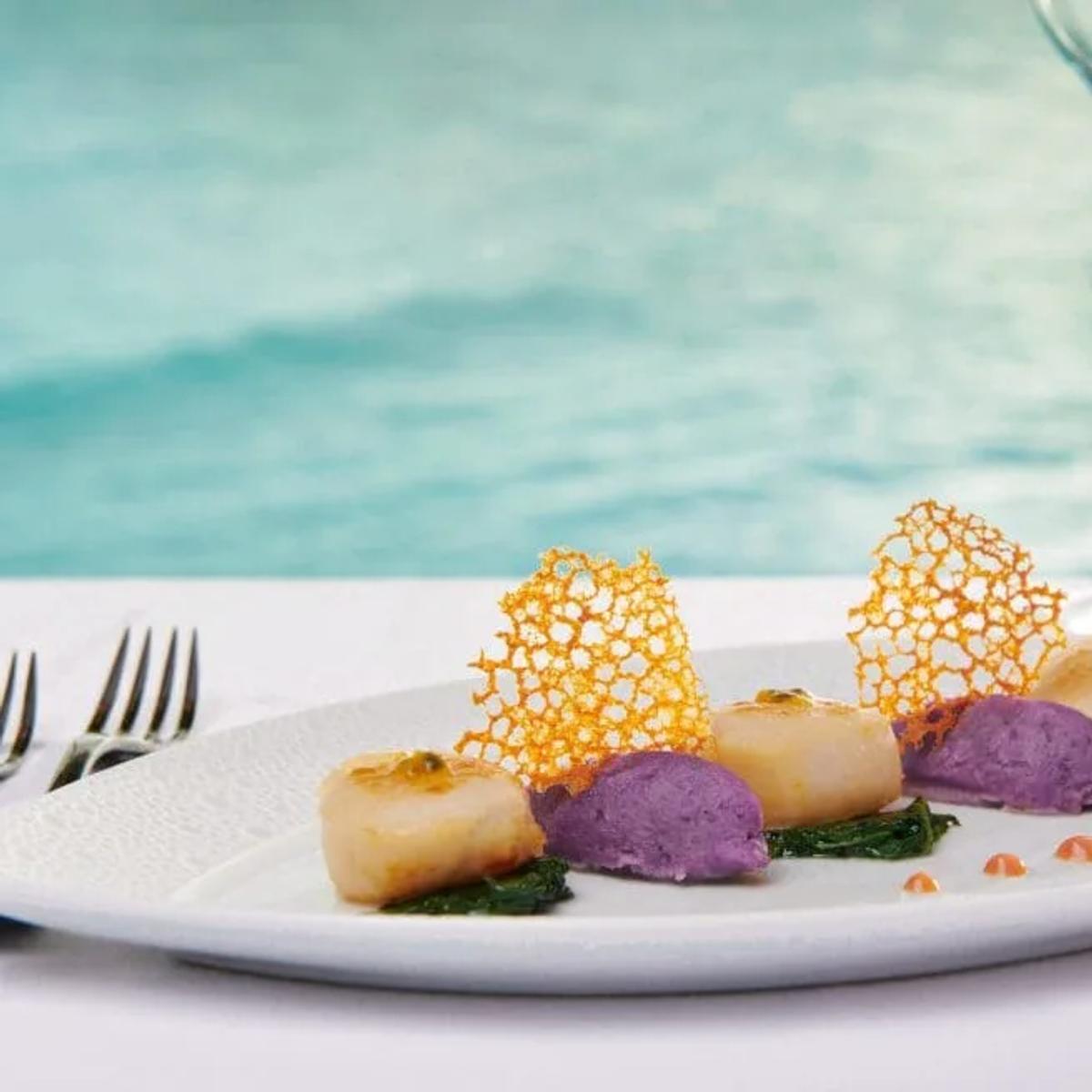 A plated seafood dish with scallops and purple potato purée, two glasses of white wine and the ocean behind