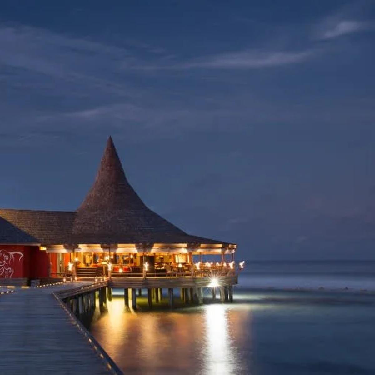 Baan Huraa's teak pavilion lit at dusk, built on stilts over the lagoon