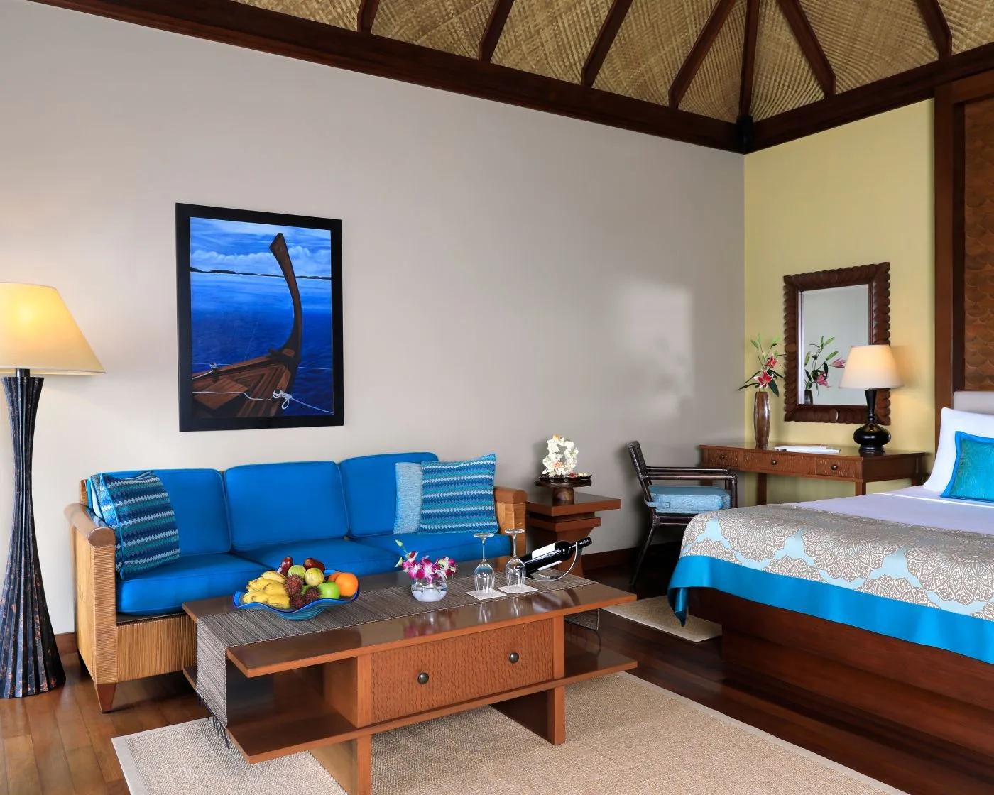 Inside a Sunrise Overwater Villa's living and bedroom area, a blue sofa beneath a framed dhoni painting