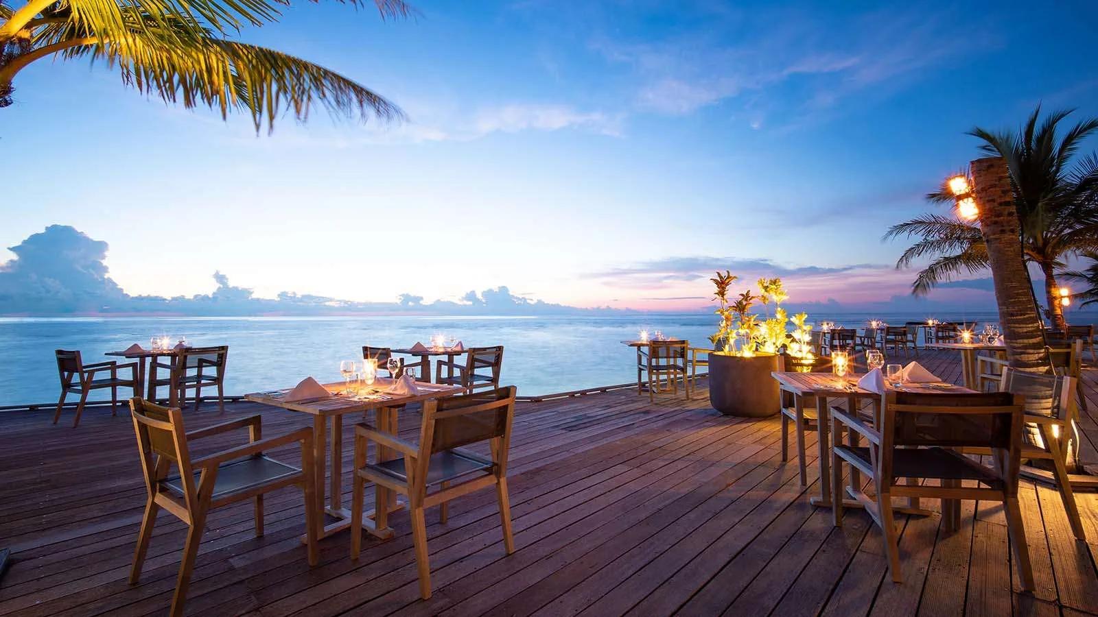 Falhu Restaurant's terrace at dusk, tables set beneath palm trees along the water's edge