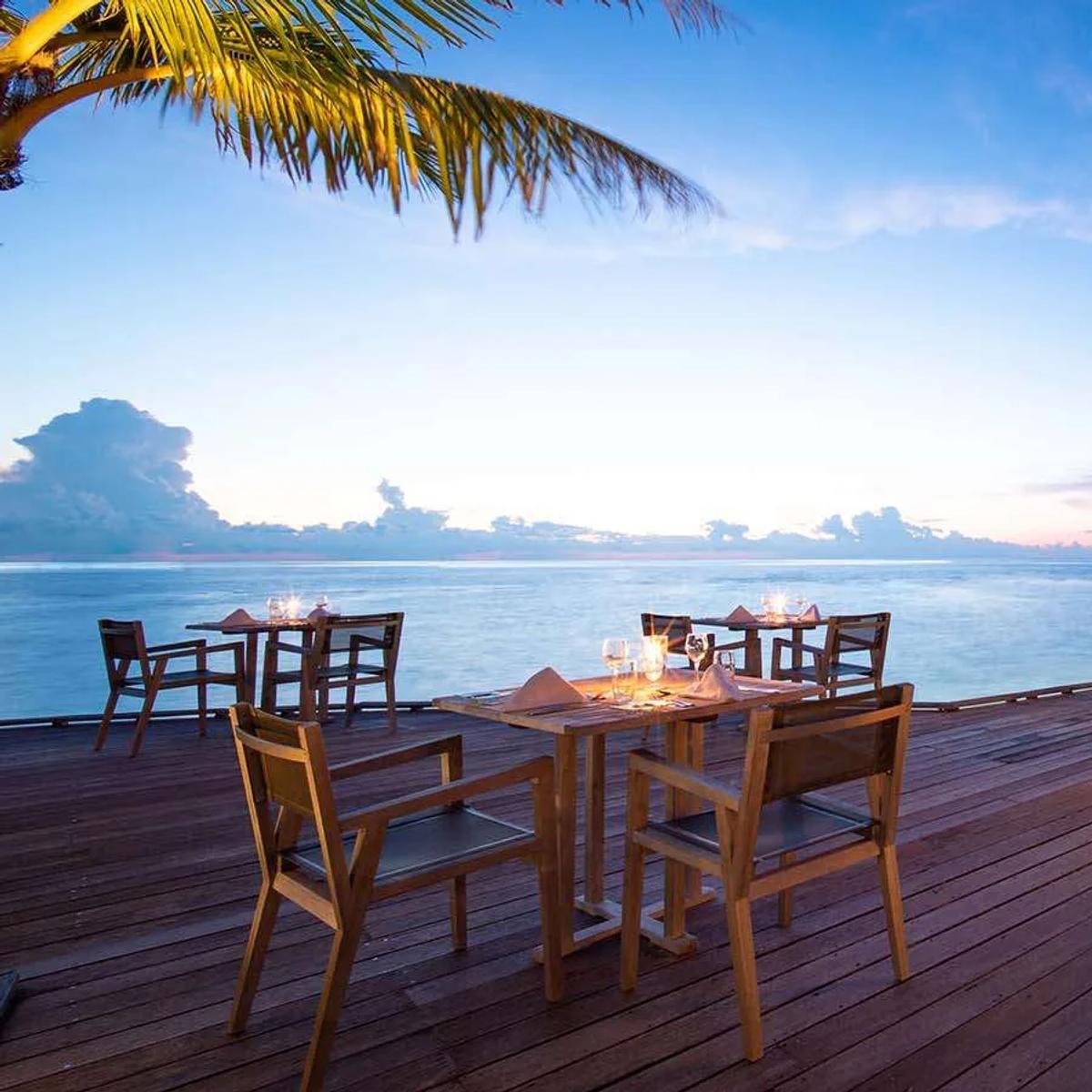 Falhu Restaurant's terrace at dusk, tables set beneath palm trees along the water's edge