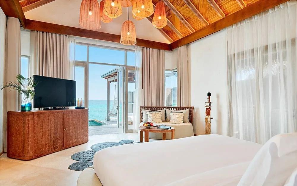 Overwater villa bedroom with a rattan sofa, wood-beamed ceiling, and glass doors opening onto a private deck over the lagoon