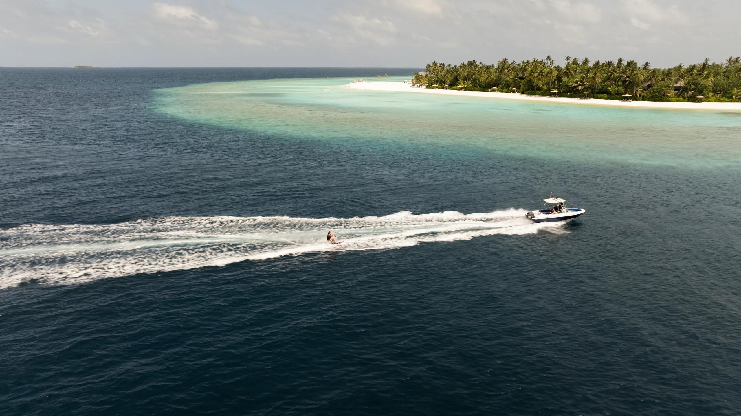 Aerial view of a speedboat towing a water-skier past Constance Halaveli's beach and reef edge, a second small island visible on the horizon