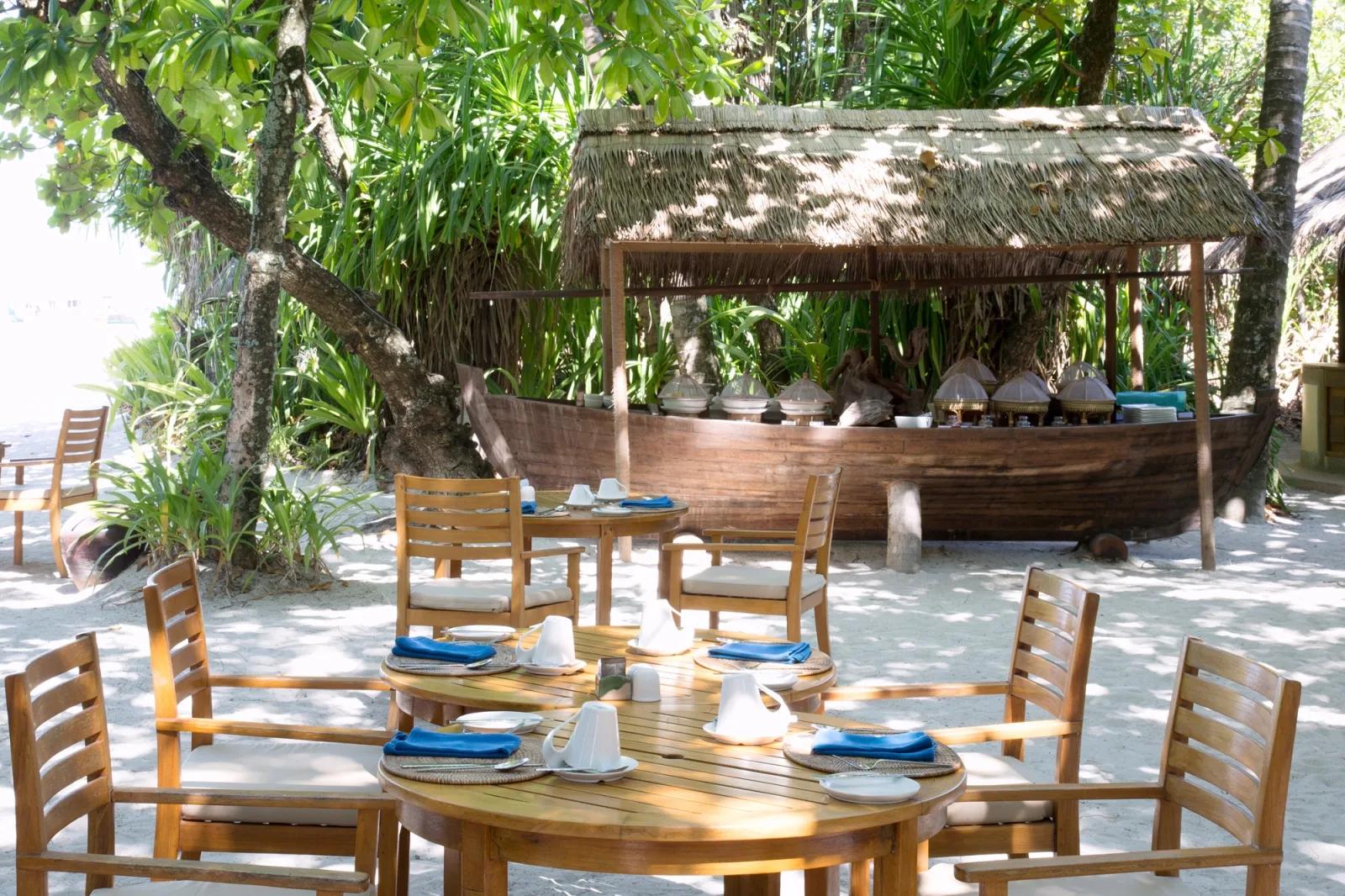 An outdoor breakfast buffet laid out on a traditional wooden boat display, tables set beneath trees on the beach