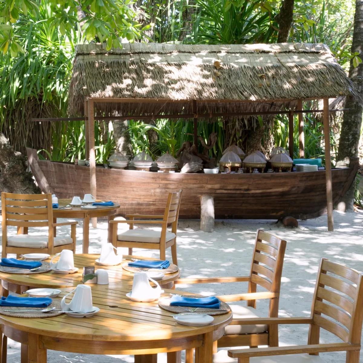 An outdoor breakfast buffet laid out on a traditional wooden boat display, tables set beneath trees on the beach