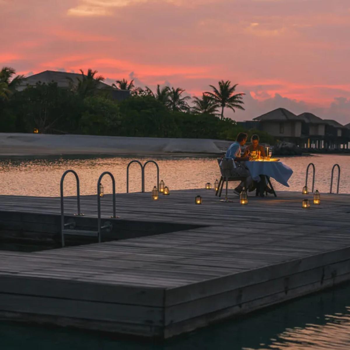 A private table set for two on an overwater pontoon at dusk, part of the resort's bespoke dining programme