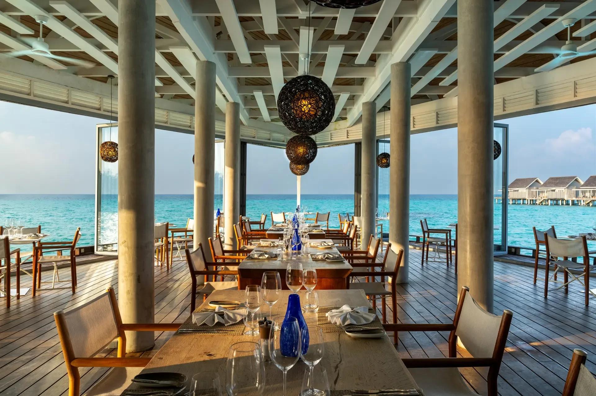 The Reef, Kuramathi's waterfront restaurant, set tables facing the open lagoon with a row of overwater villas visible in the distance at dusk