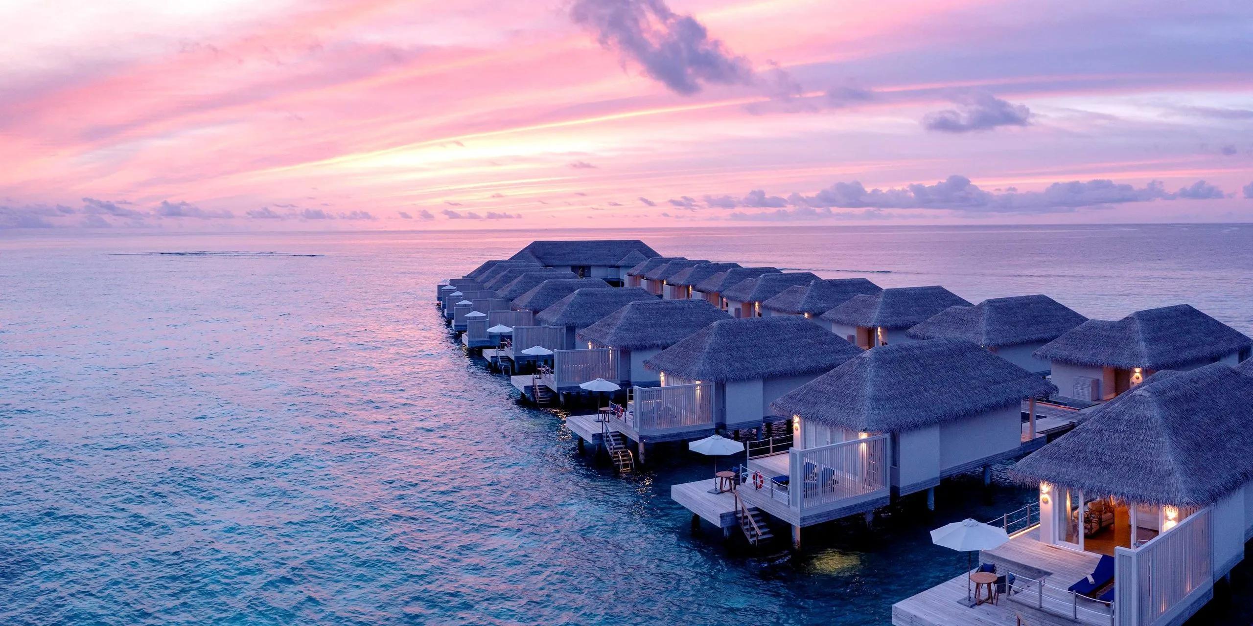 A row of overwater Sunset Water Villas at Baglioni Maldives under a pink and purple dusk sky