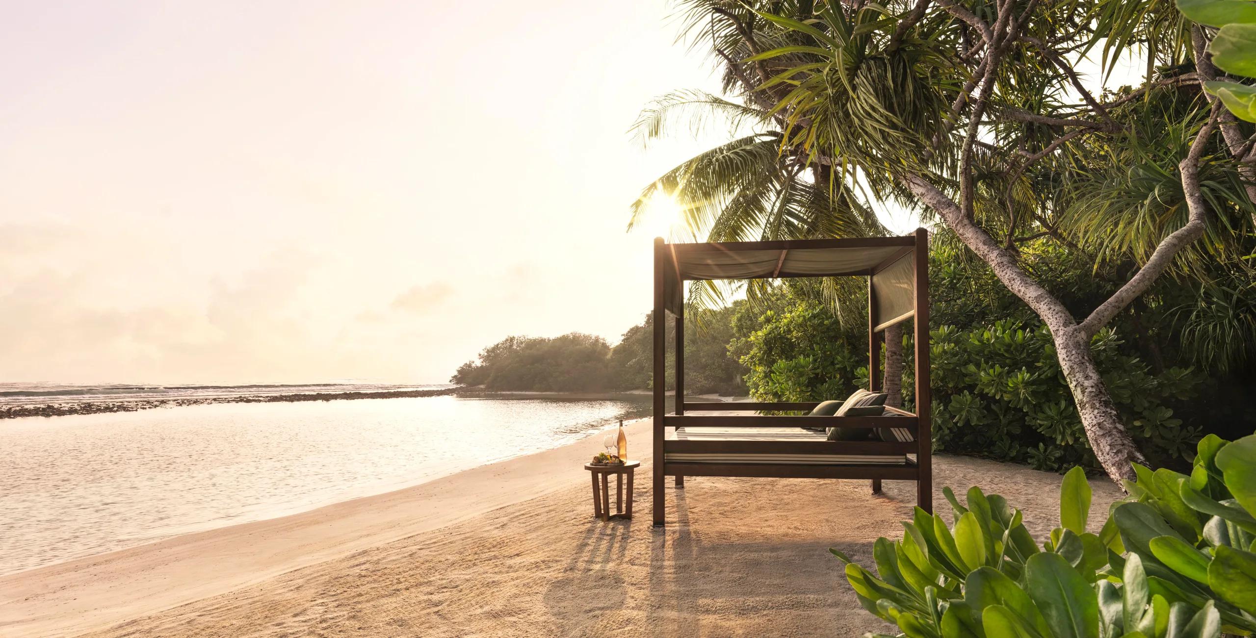 A canopied daybed set alone on a quiet stretch of beach at sunrise, with wine and snacks laid out on a side table.