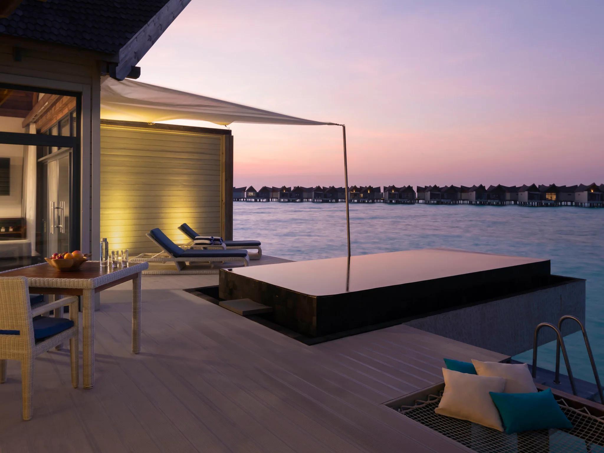 A private dining table set for two on a villa's overwater deck at dusk, the plunge pool alongside and a row of villas silhouetted beyond