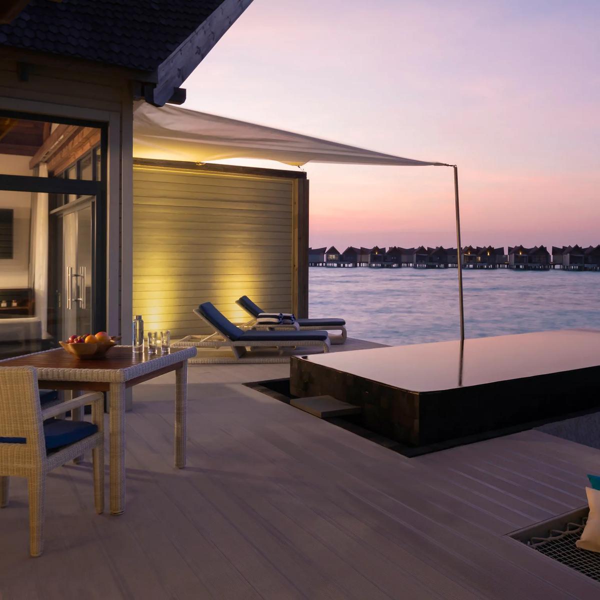 A private dining table set for two on a villa's overwater deck at dusk, the plunge pool alongside and a row of villas silhouetted beyond