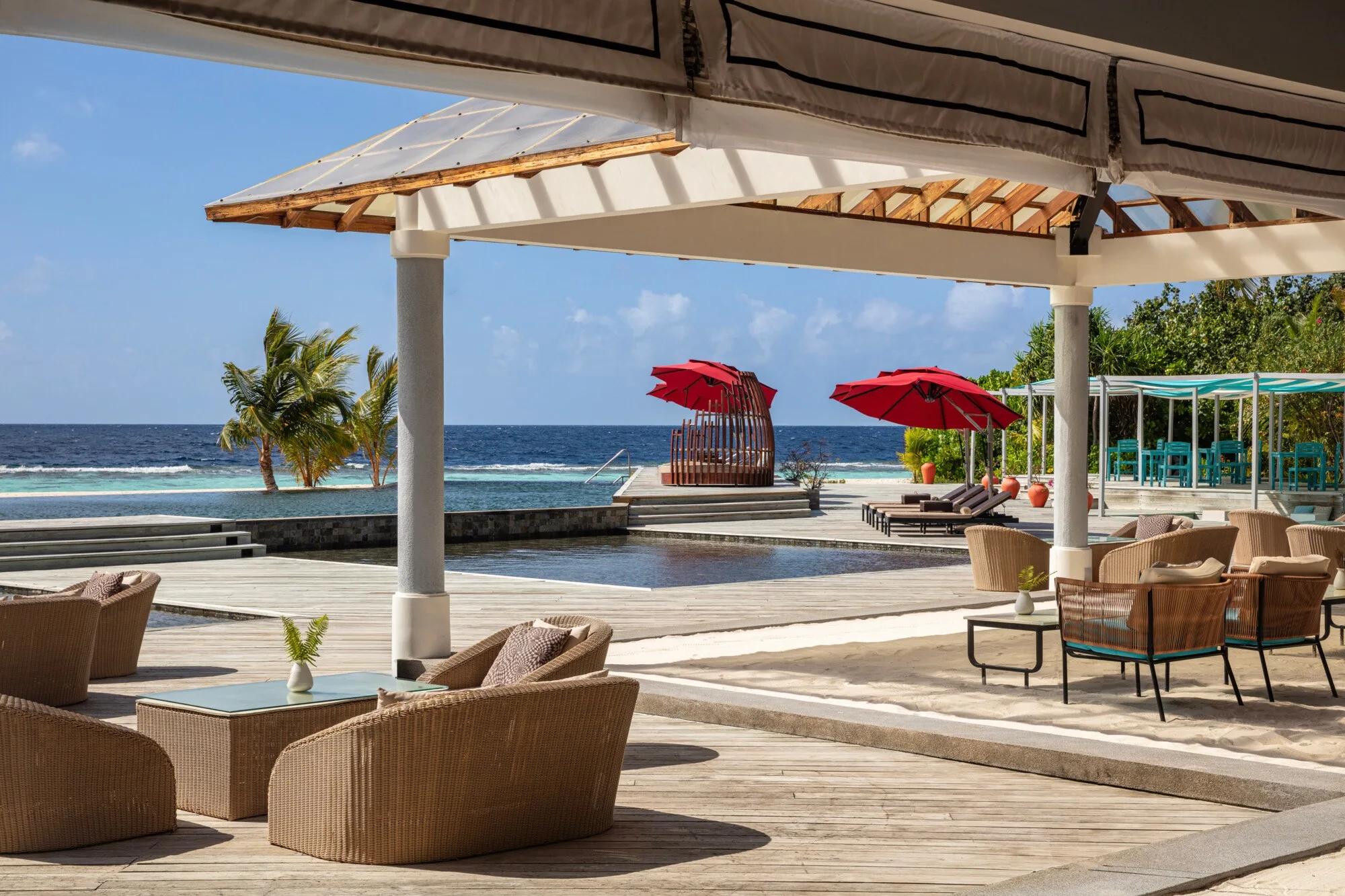 An elevated pool deck with red umbrellas, a woven daybed and loungers, the open sea visible beyond the infinity-edge pool