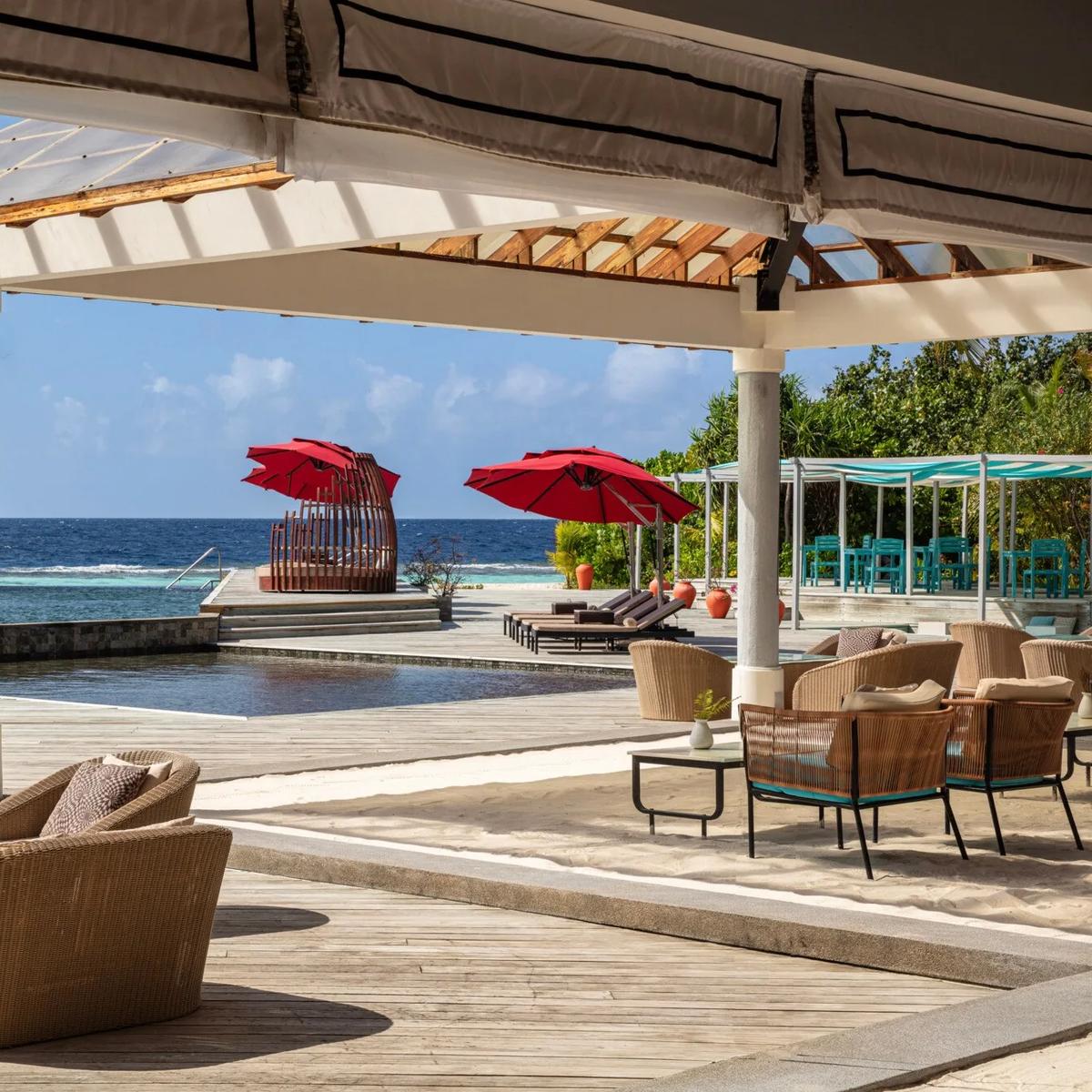 An elevated pool deck with red umbrellas, a woven daybed and loungers, the open sea visible beyond the infinity-edge pool