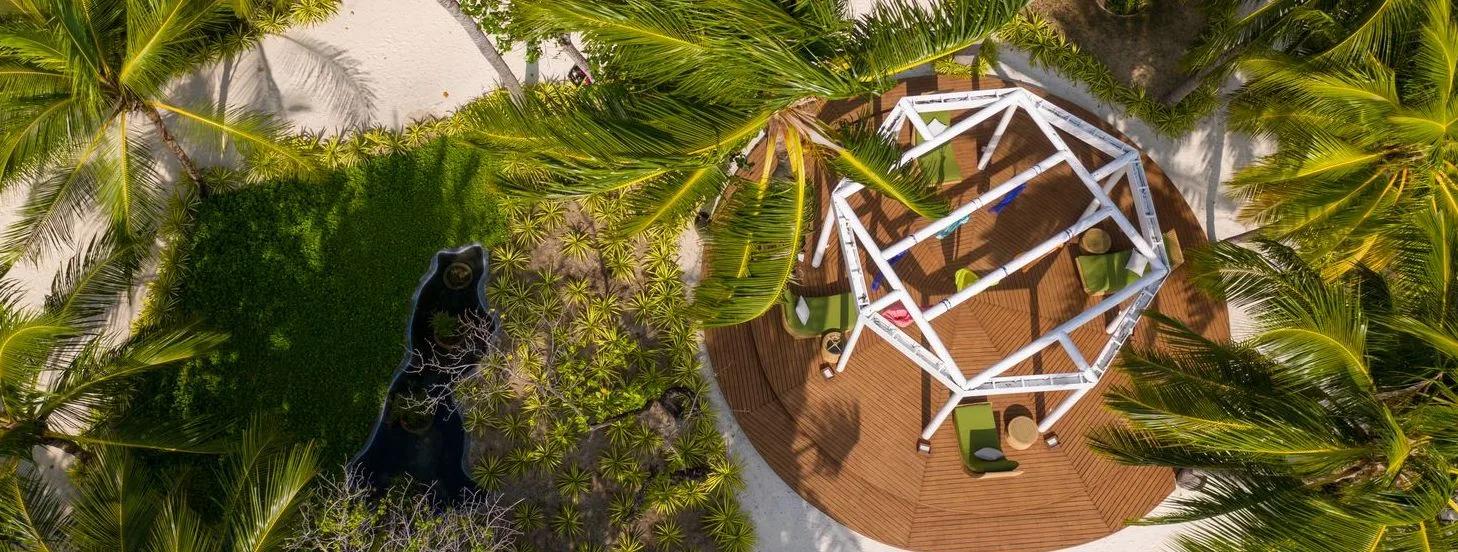 Aerial view of a circular wooden wellness deck with a geometric frame installation, shaded by palm trees
