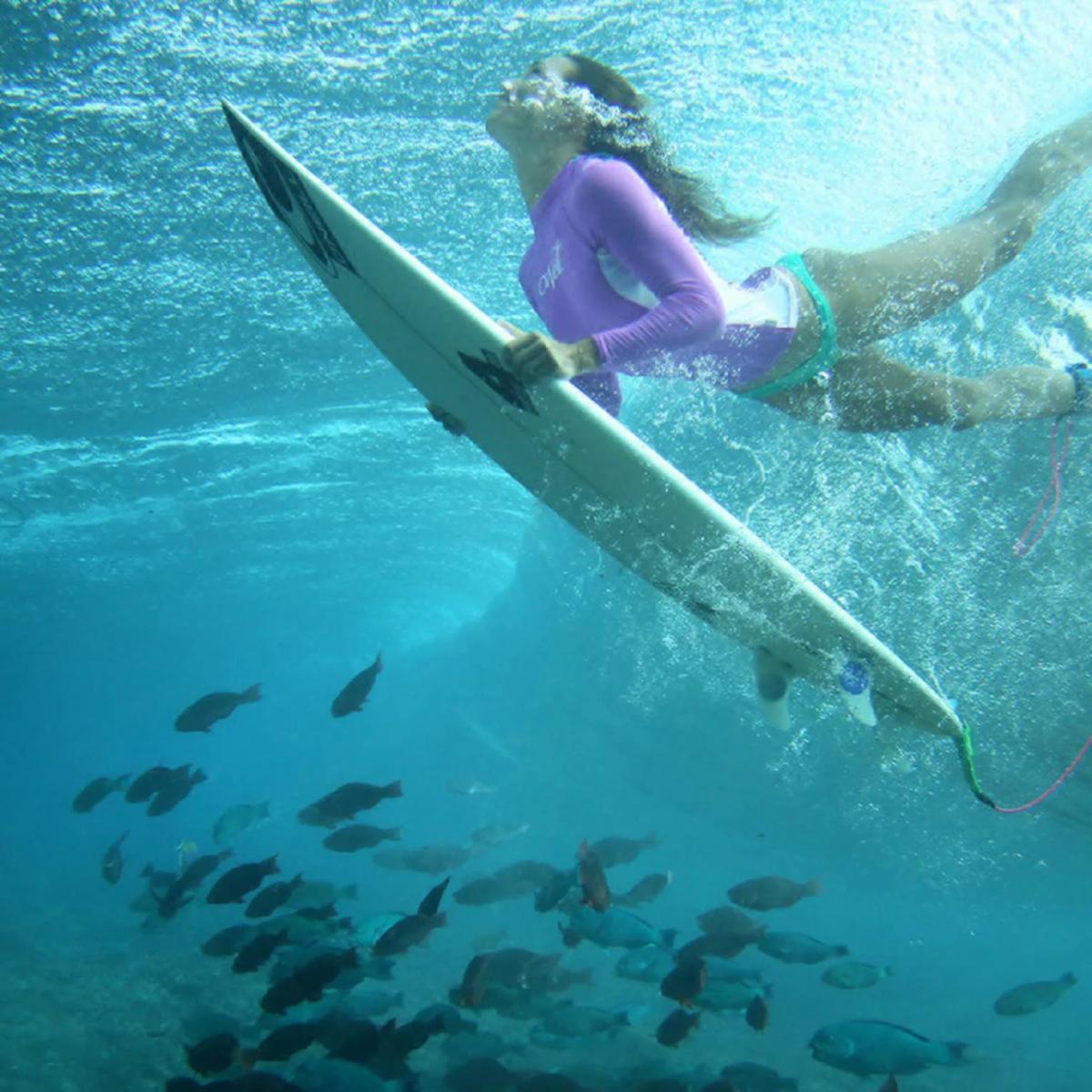 A surfer diving beneath a wave with reef fish schooling below, seen from underwater