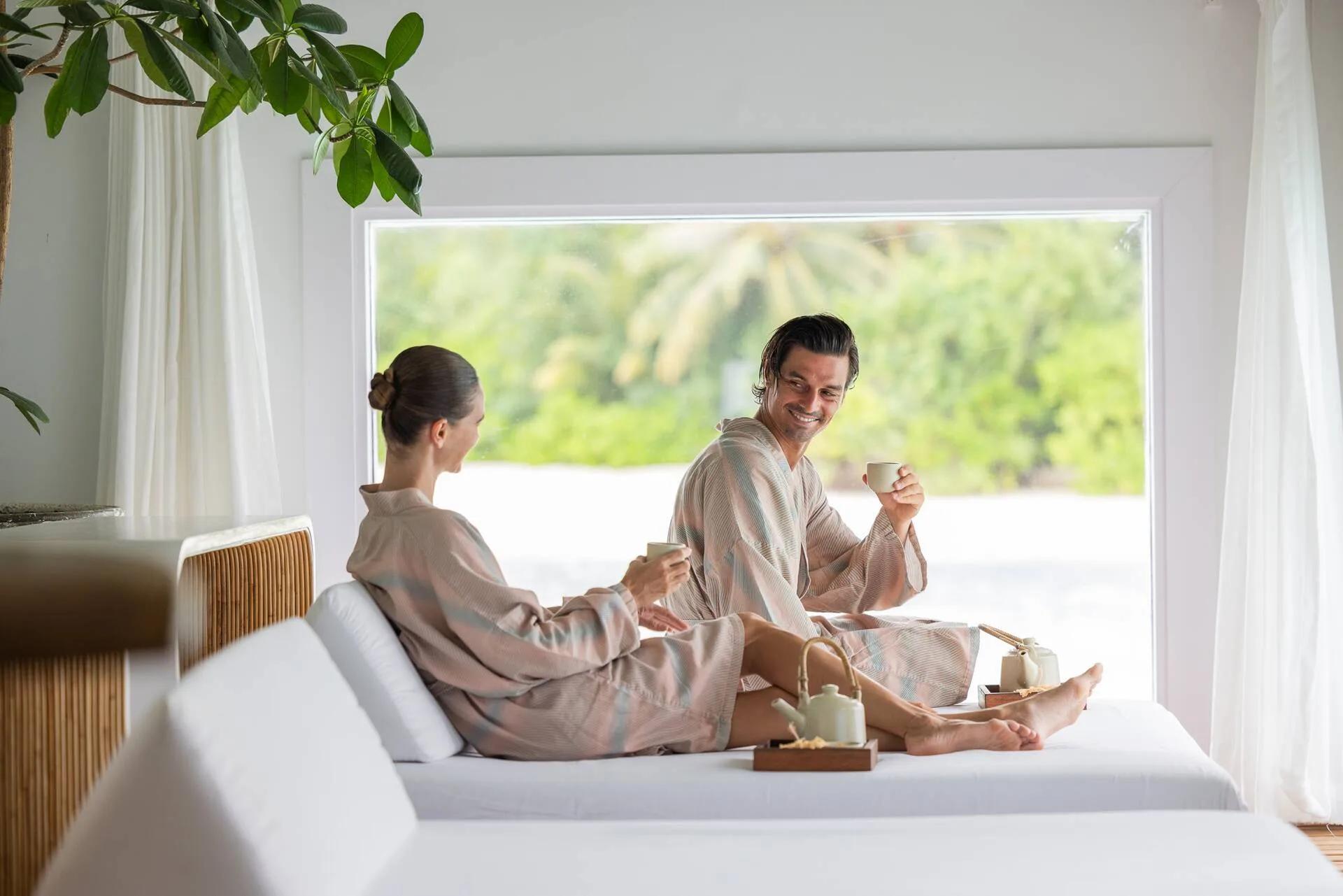 A couple in robes drinking tea on a daybed at The Standard Spa, the beach visible through an open doorway behind them