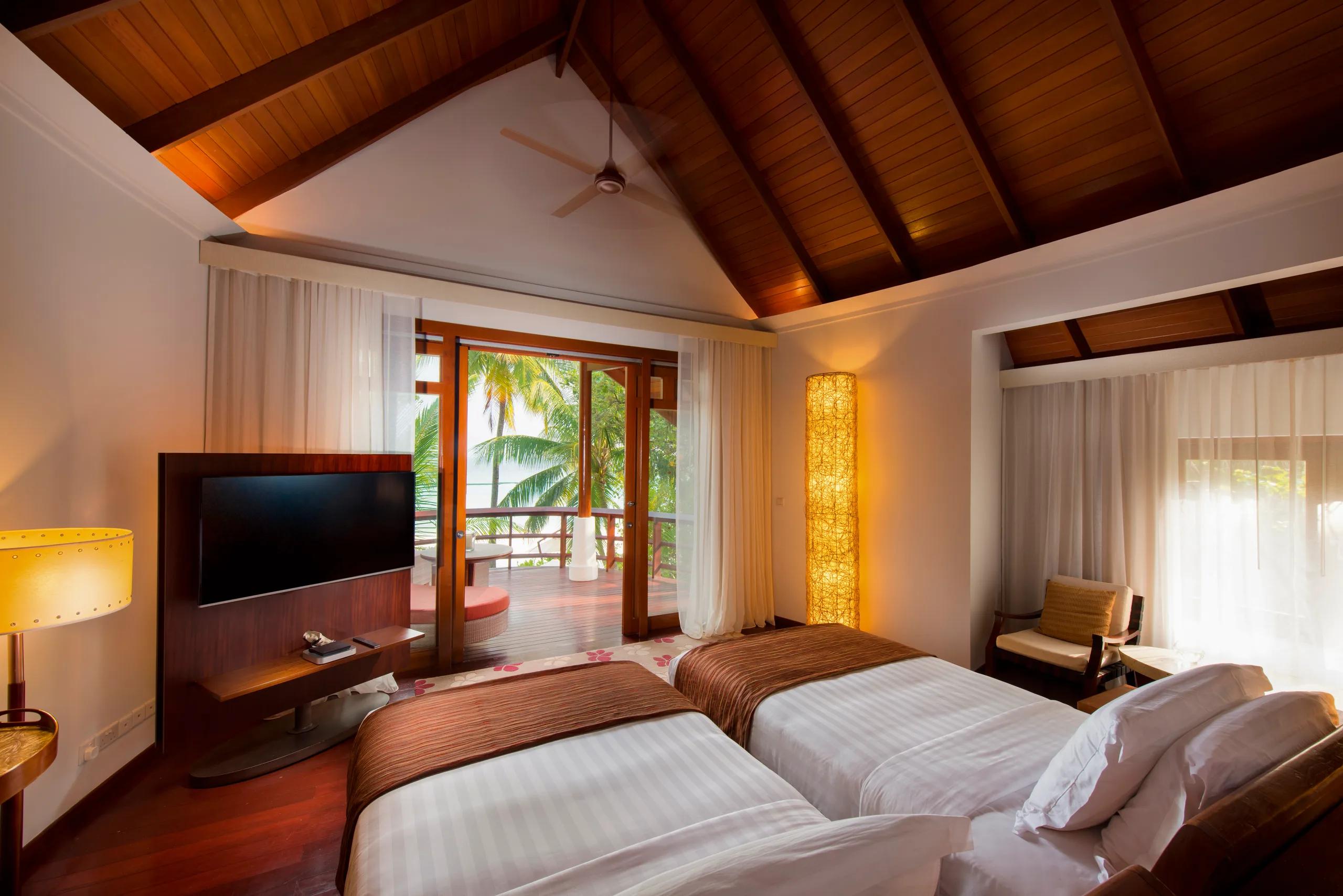 A Double Storey Beach Villa bedroom with twin beds and a wall-mounted TV, glass doors open to a furnished terrace with a sea view beyond
