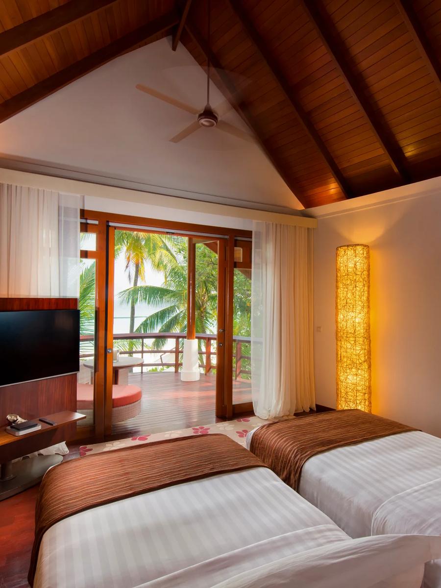 A Double Storey Beach Villa bedroom with twin beds and a wall-mounted TV, glass doors open to a furnished terrace with a sea view beyond