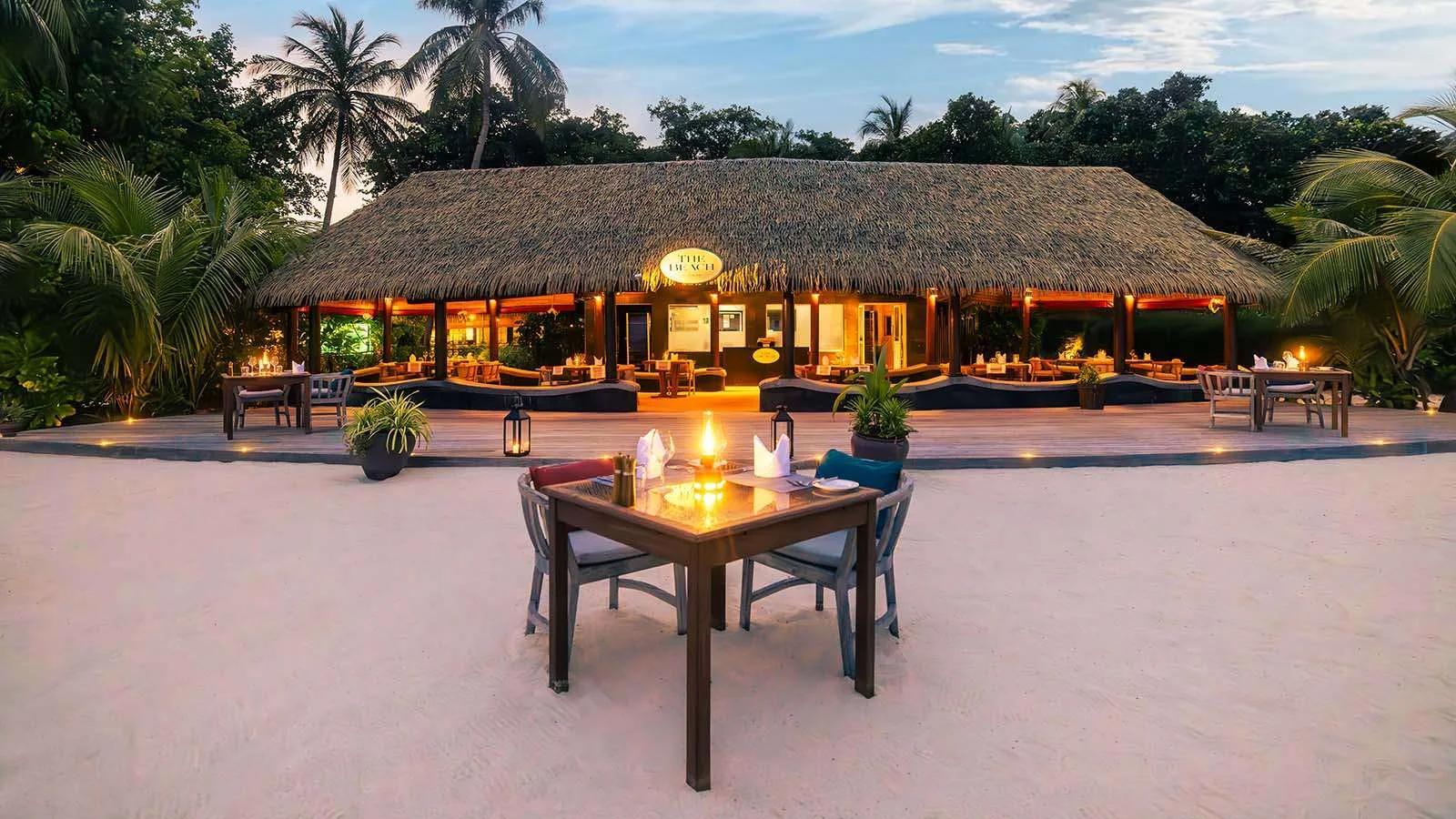 The Beach restaurant's open-air tables lit at dusk under palm trees, ocean beyond