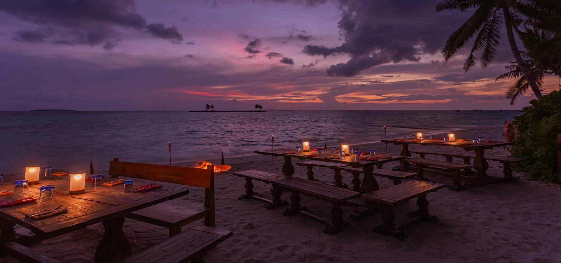 BBQ Shak's beach dinner tables lit by lanterns and torches at dusk, a small island silhouetted on the horizon