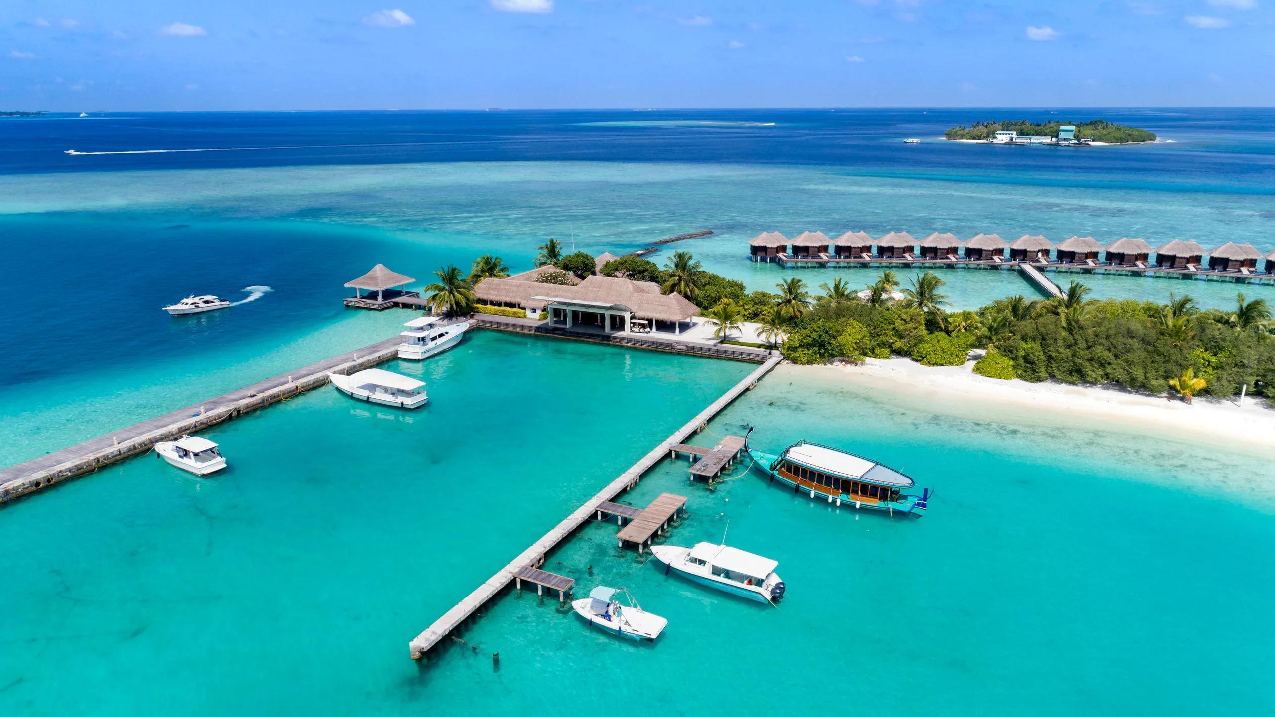 Aerial view of the resort's marina, with boats moored along a jetty and the overwater villa row visible beyond the trees