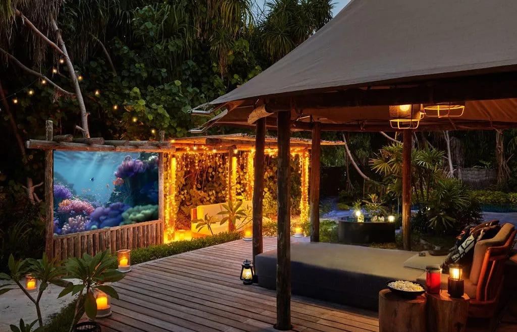 Jungle Cinema's outdoor screen showing an underwater reef scene, with string lights and lounge seating set beneath a thatched canopy at night
