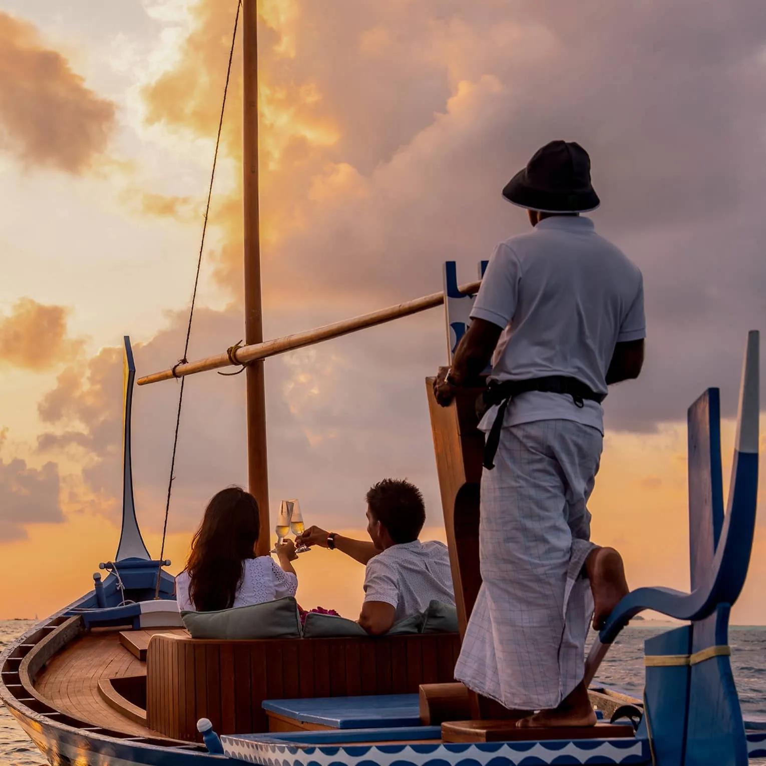A couple toasting champagne aboard a traditional dhoni at sunset, with a crew member steering from the bow