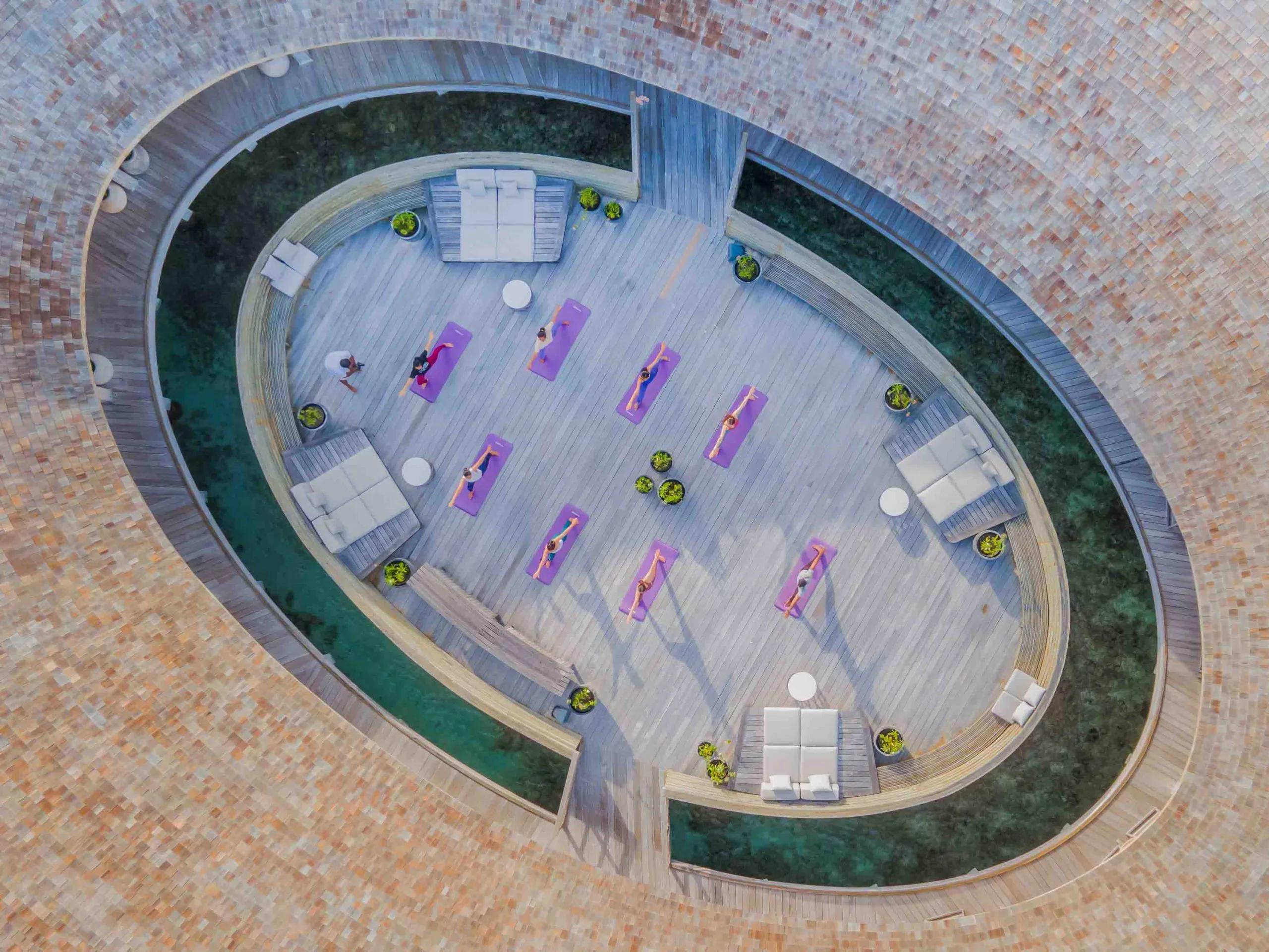 An aerial view of a yoga class on a round, water-ringed deck, mats laid out in rows