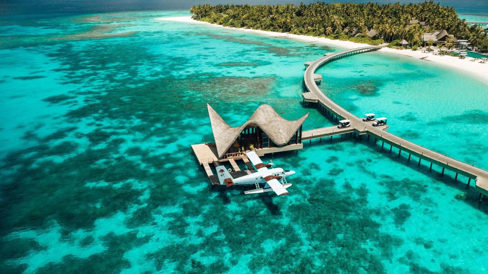 A seaplane docked beside JOALI Maldives' overwater lounge, the jetty curving back to Muravandhoo Island's beach with reef visible through the clear lagoon