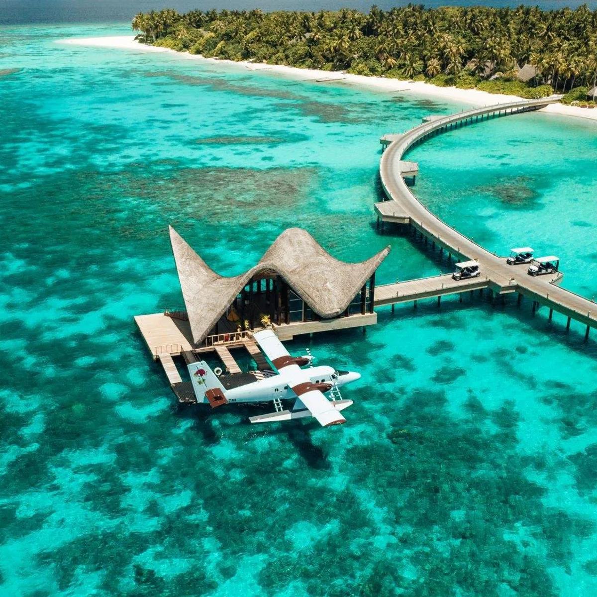 A seaplane docked beside JOALI Maldives' overwater lounge, the jetty curving back to Muravandhoo Island's beach with reef visible through the clear lagoon