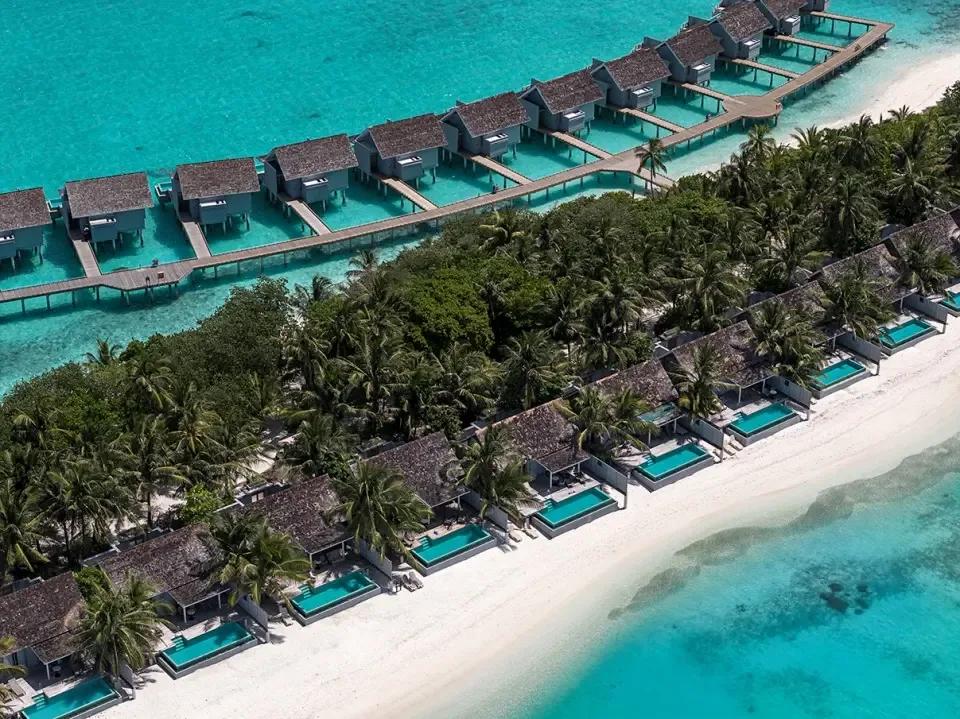 Aerial view of Kuramathi's overwater villa jetty and a row of beachfront pool villas along the shoreline, framed by palm trees
