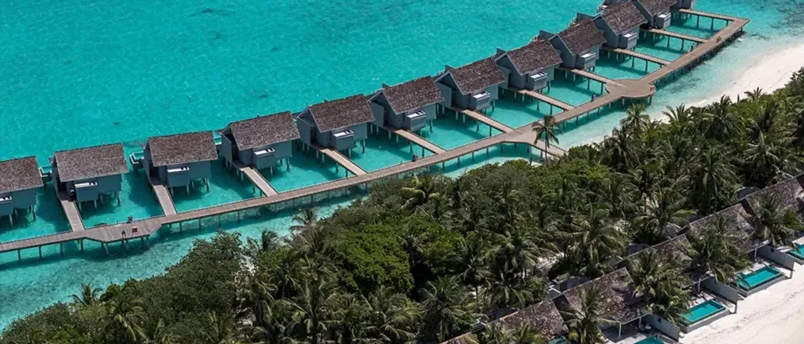 Aerial view of Kuramathi's overwater villa jetty and a row of beachfront pool villas along the shoreline, framed by palm trees