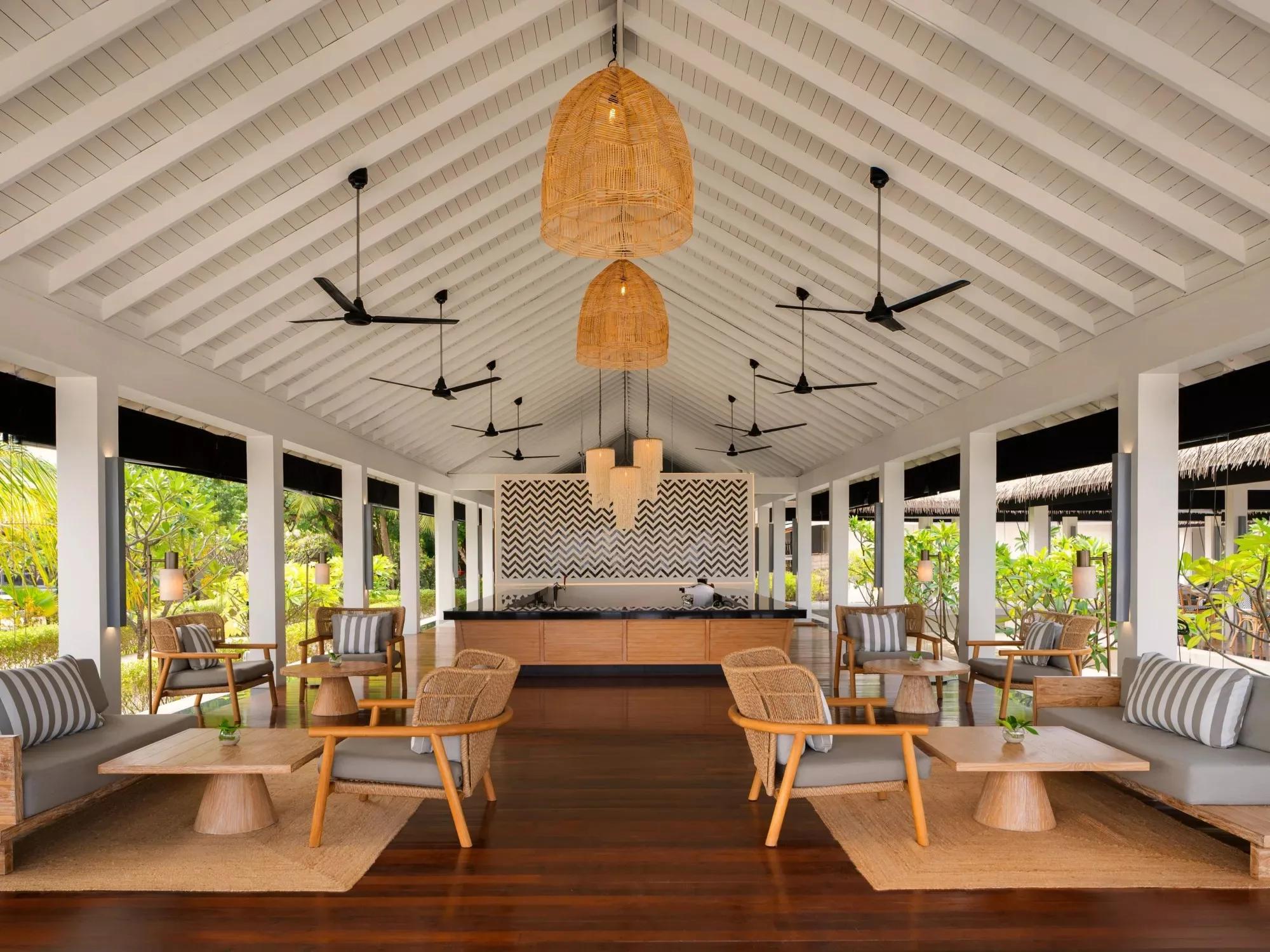 An open-air lounge and bar area at Noku Maldives, rattan seating beneath a vaulted thatched ceiling