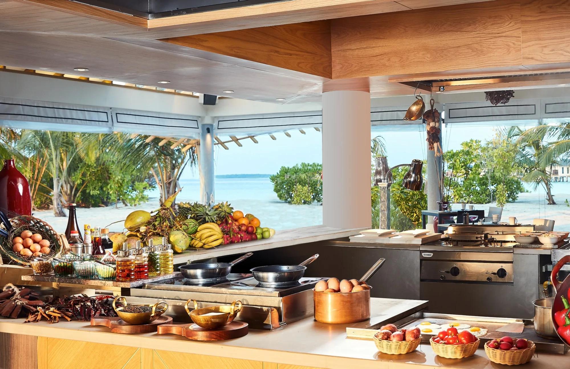 REEF's open-kitchen buffet counter laid with fresh fruit, eggs, cured meats and condiments, the lagoon and palms visible through the open-air pavilion behind