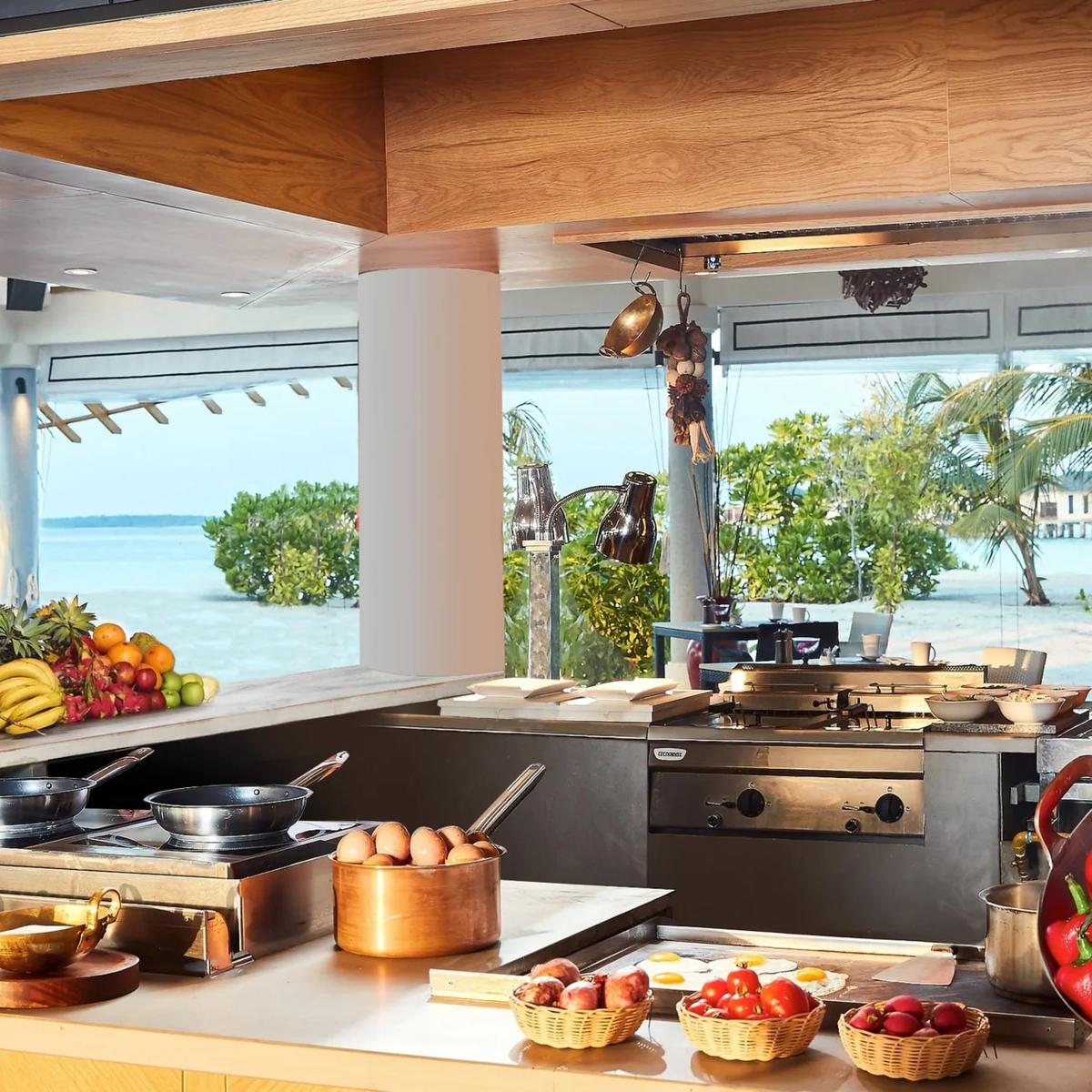 REEF's open-kitchen buffet counter laid with fresh fruit, eggs, cured meats and condiments, the lagoon and palms visible through the open-air pavilion behind