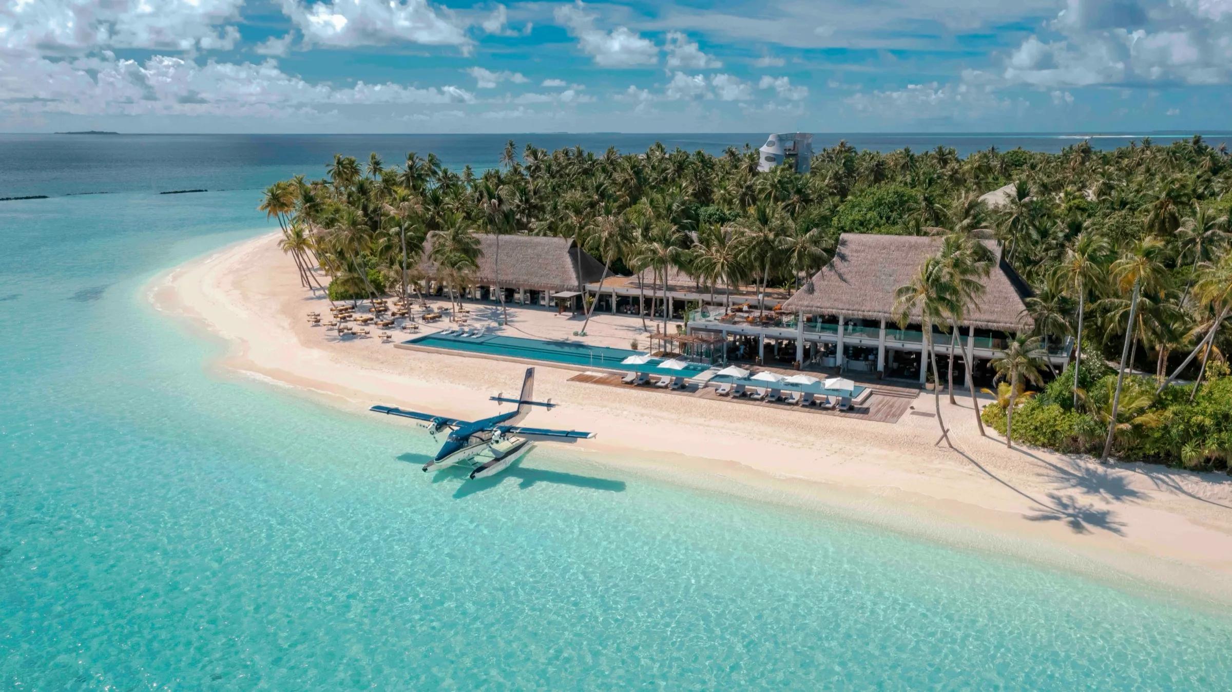 A seaplane moored on the sand beside Velaa's beachfront restaurant and pool deck, palm trees framing the arrival point