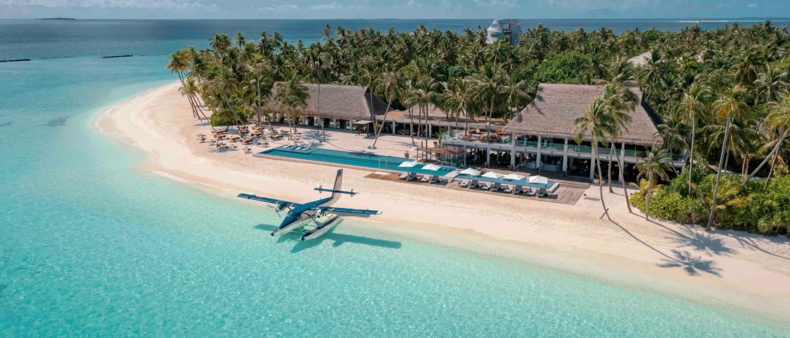 A seaplane moored on the sand beside Velaa's beachfront restaurant and pool deck, palm trees framing the arrival point