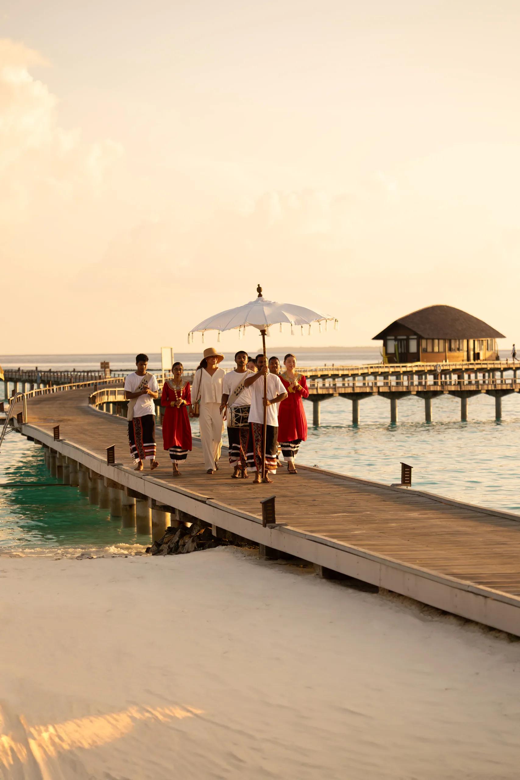 A staff member in traditional dress leading a parasol-shaded welcome walk along an overwater jetty