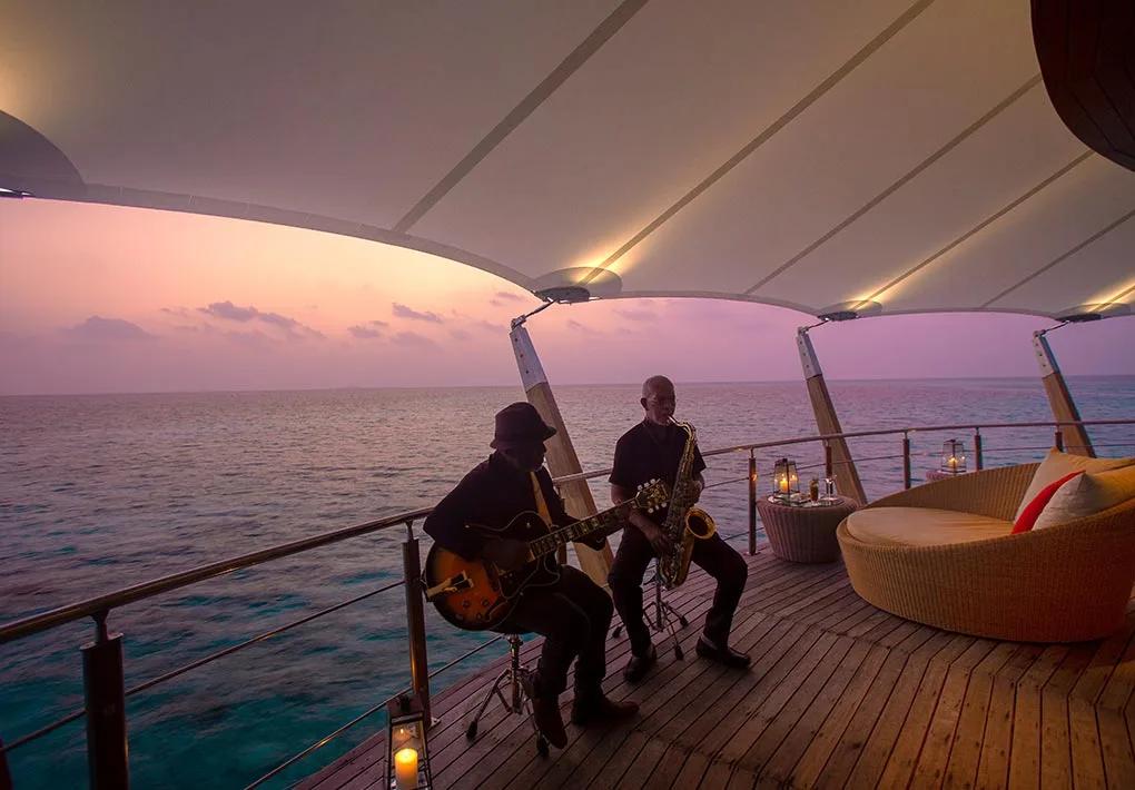 Two musicians playing guitar and saxophone on an overwater deck under a sail-shaped canopy as the sun sets over the ocean